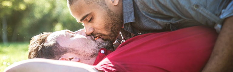 Two men lie on the sunny grass of a park, kissing passionately.
