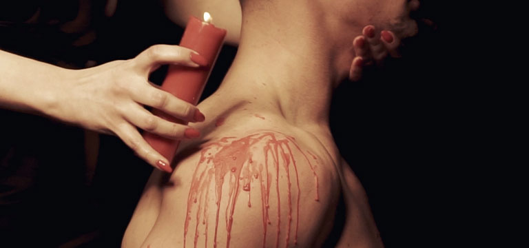 A woman uses a red drip candle to drip hot wax over her lover's muscular shoulders.