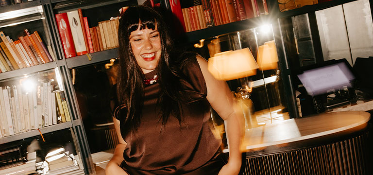 Laura Roscioli reclines against a counter in front of a bookshelf at a sexy book club event.